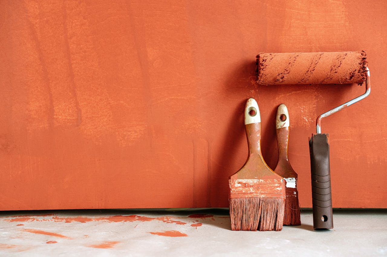 about-06 Close-up of paint rollers and brushes with orange paint against a painted wall.